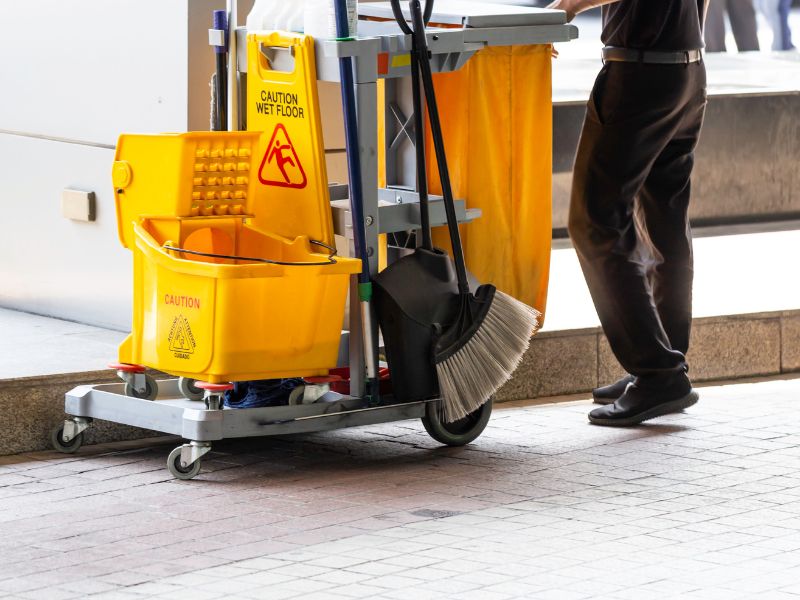Janitor cart with a mop bucket, caution sign, and cleaning supplies on a tiled floor. A person in dark clothing is partially visible, standing next to the cart.