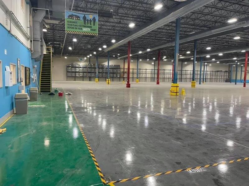 A large, empty warehouse with shiny floors and a high ceiling. Yellow bollards and red support beams are visible throughout the space. Shelving units line the back wall, and a blue wall with bulletin boards is on the left.