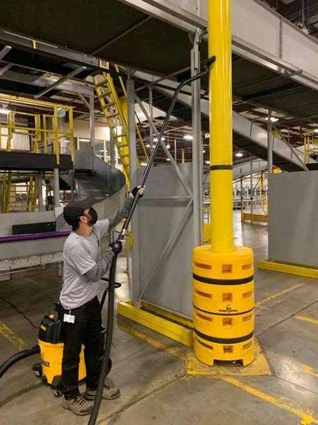 A person in work attire uses a vacuum device to clean a high spot on a yellow post in an industrial setting. Conveyor belts and metal structures are visible in the background. The area appears to be a factory or warehouse.