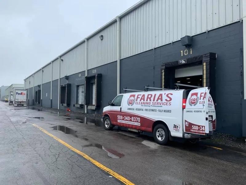 A white van with "Faria's Cleaning Services" branding is parked outside a large industrial warehouse building with gray walls. A FedEx truck is visible further down the wet pavement on a rainy day.