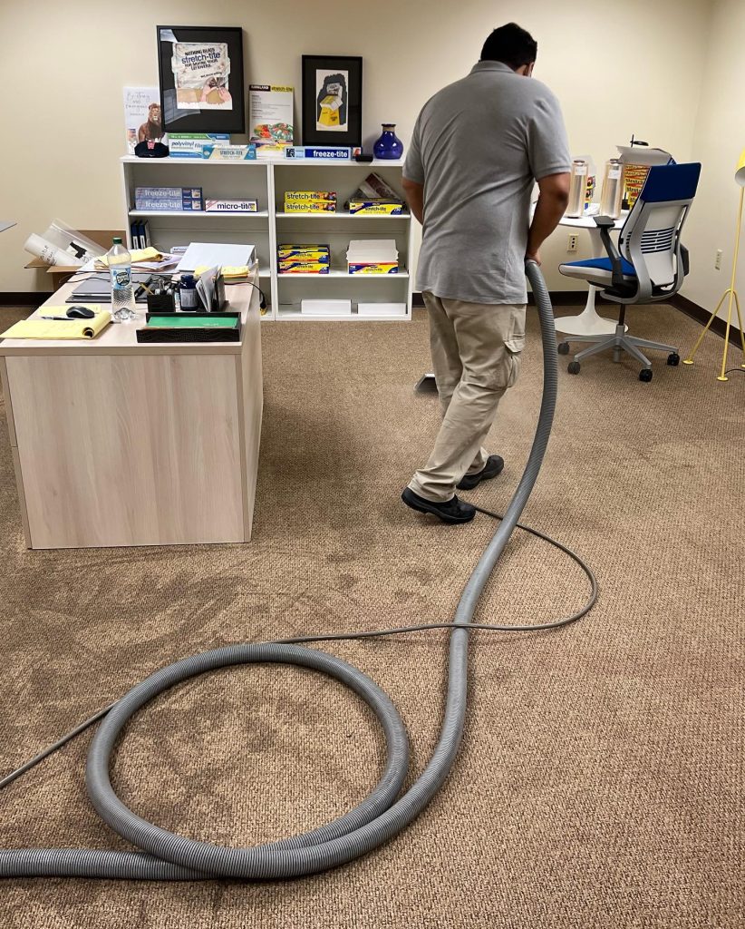 A person in a gray shirt vacuums a beige carpeted office with a large hose. The office has a desk, chair, bookshelves, and various office supplies. Framed posters and vases are visible on the shelves.