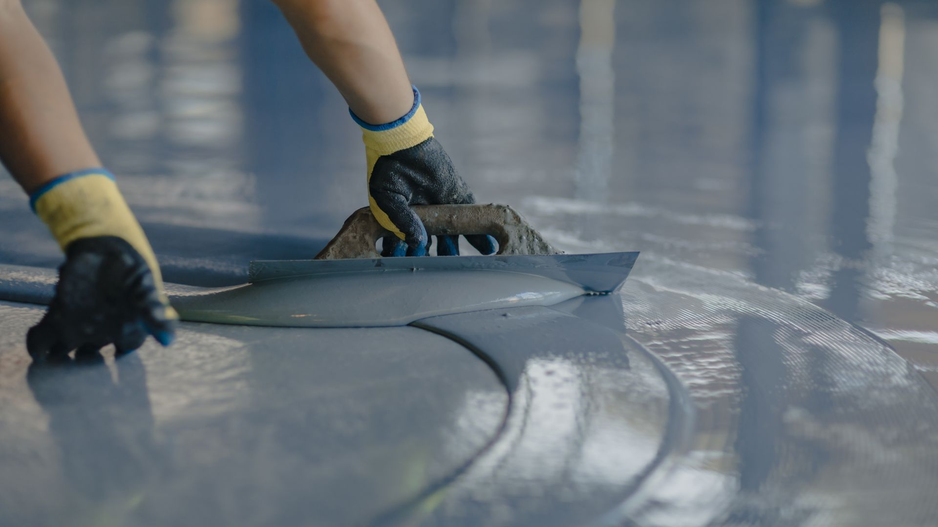 A person wearing gloves is using a trowel to smooth and spread a glossy blue-gray epoxy resin on a floor. The surface is wet and reflective, indicating ongoing work.