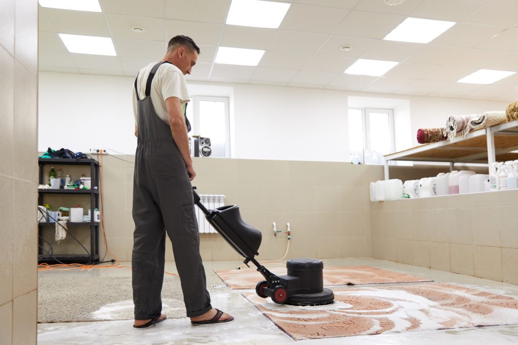 A person wearing overalls operates a floor cleaning machine on a rug inside a well-lit room. Shelves with cleaning supplies and rolled-up rugs are in the background.