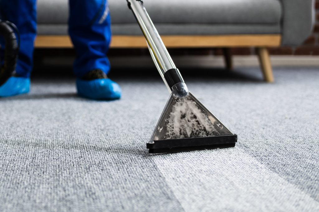 A person wearing blue shoe covers uses a carpet cleaning machine on a gray carpet in a room with a sofa in the background. The left part of the carpet looks cleaner than the right.