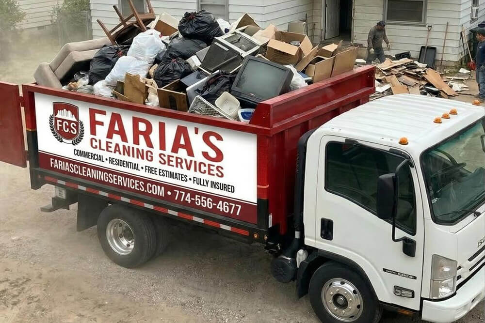 A large Faria’s Cleaning Services truck is filled with junk, including old electronics, bags, and furniture, parked beside a house where people are clearing debris.