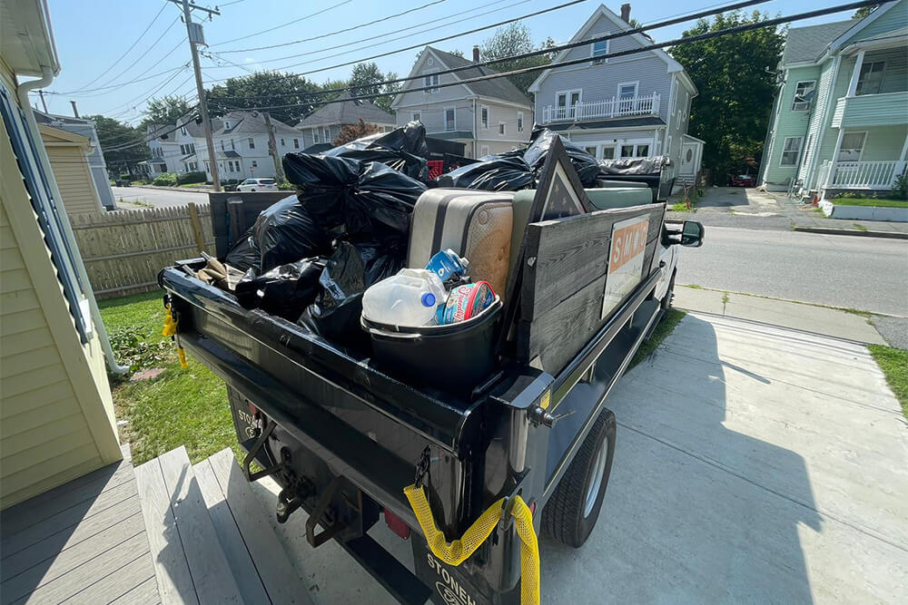 A truck loaded with black garbage bags, cardboard, and recyclables is parked in a residential driveway with houses and a street visible in the background on a sunny day.